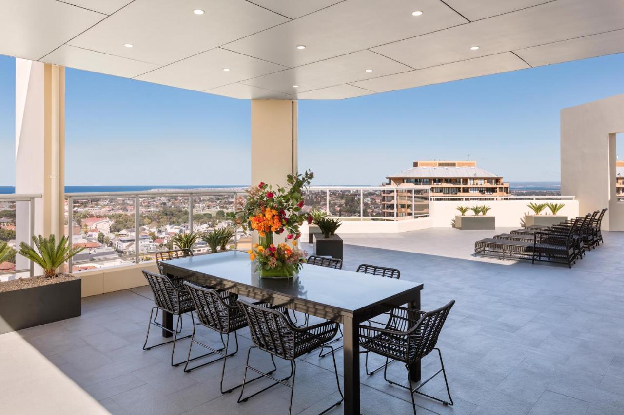 Meriton Suites Bondi Junction Sidney Exterior foto The photo shows a stylish outdoor terrace or balcony area, featuring a long dining table surrounded by several black wire chairs. The table has a centerpiece of vibrant flowers. The space is well-lit, with a clear view of the horizon in the backgroun