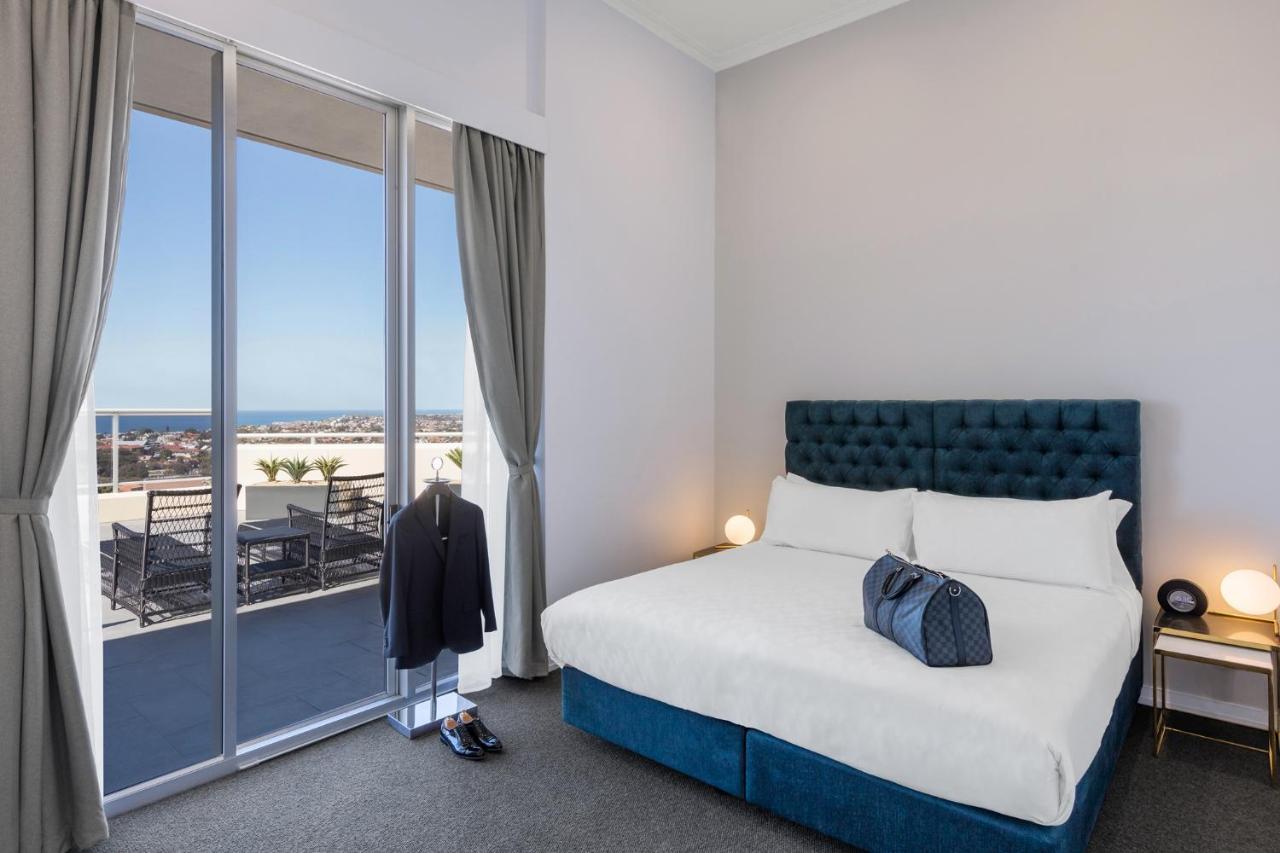 Meriton Suites Bondi Junction Sidney Exterior foto The photo shows a stylish bedroom featuring a large bed with a tufted blue headboard. There's a neatly organized outfit consisting of a suit hanging on a stand, along with shoes placed on the floor. The room has large windows that provide a view of t