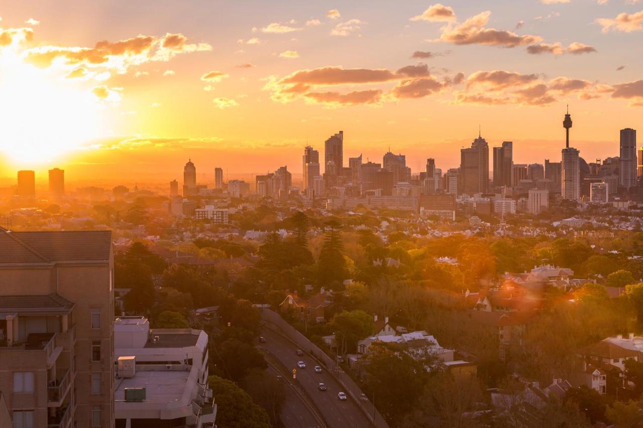 Meriton Suites Bondi Junction Sidney Exterior foto The photo shows a panoramic view of a city skyline at sunset. The sun is low on the horizon, casting a warm golden light across the buildings. Tall skyscrapers are silhouetted against the colorful sky, which features hues of orange, pink, and yellow,
