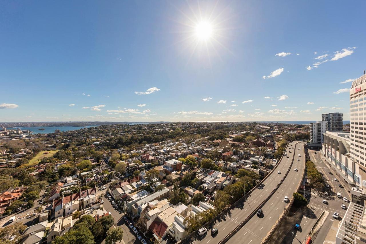 Meriton Suites Bondi Junction Sidney Exterior foto The photo shows a panoramic view of a city landscape under a clear blue sky. In the foreground, there is a road with vehicles traveling along it, bordered by trees and buildings. In the background, you can see a residential area with various houses, 