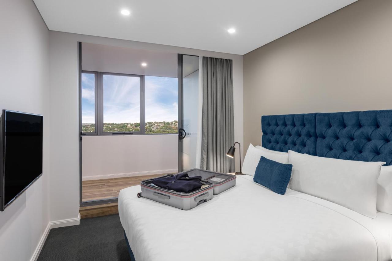 Meriton Suites Bondi Junction Sidney Exterior foto The photo shows a modern hotel room featuring a double bed with a tufted blue headboard. There is a neatly arranged suitcase placed open on the bed, indicating that someone may be unpacking. The room has light-colored walls and a window allowing natu