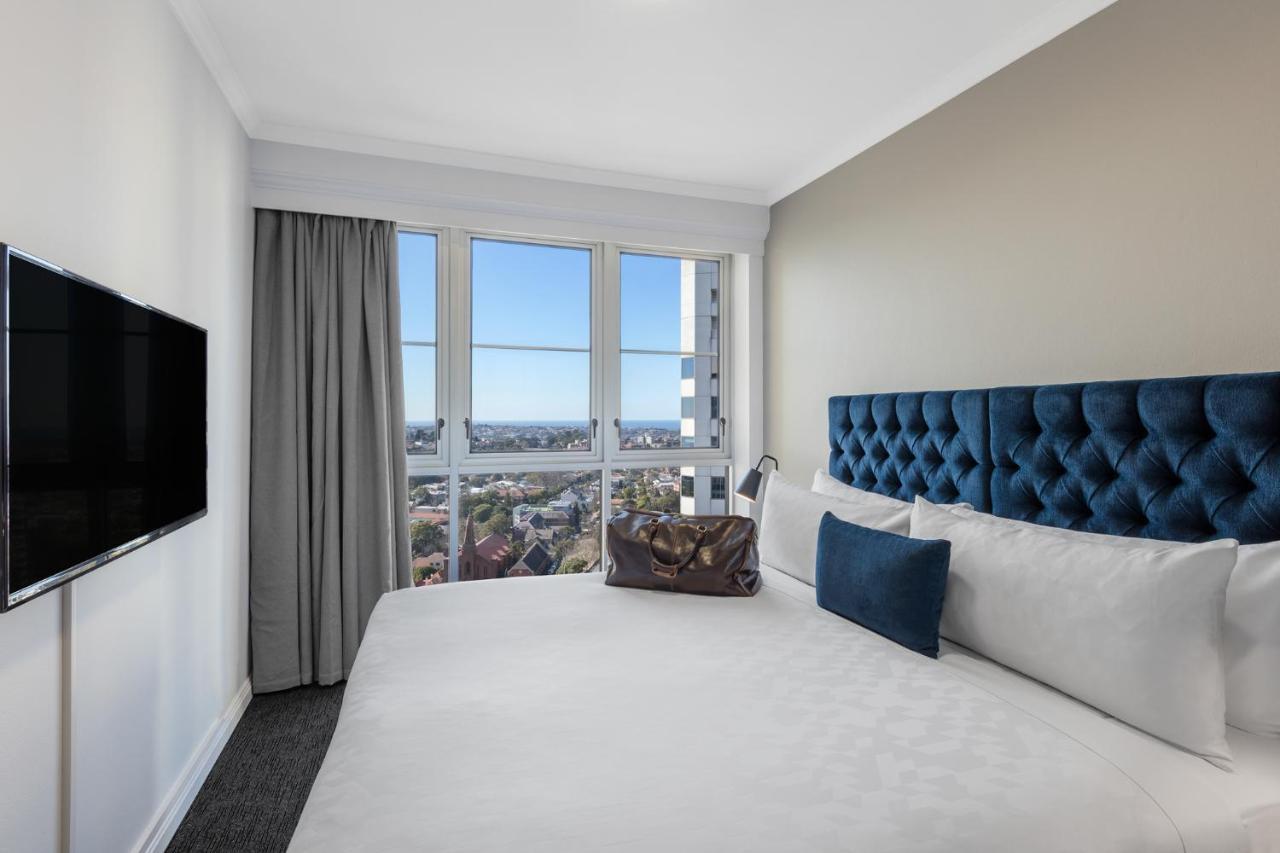 Meriton Suites Bondi Junction Sidney Exterior foto The photo shows a modern hotel room featuring a large bed with a tufted blue headboard. Beside the bed, there is a stylish bag placed on the bed. The room has large windows that provide a view of the outside, allowing natural light to fill the space.