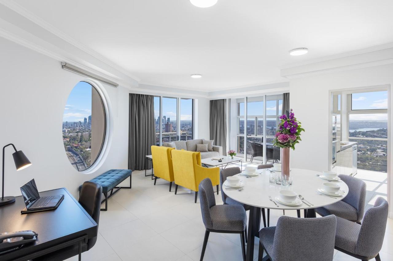 Meriton Suites Bondi Junction Sidney Exterior foto The photo shows a modern and bright interior space, likely a living room or dining area. It features large windows that offer a panoramic view of the city skyline. The furnishings include a round dining table set for several people, with plates and g