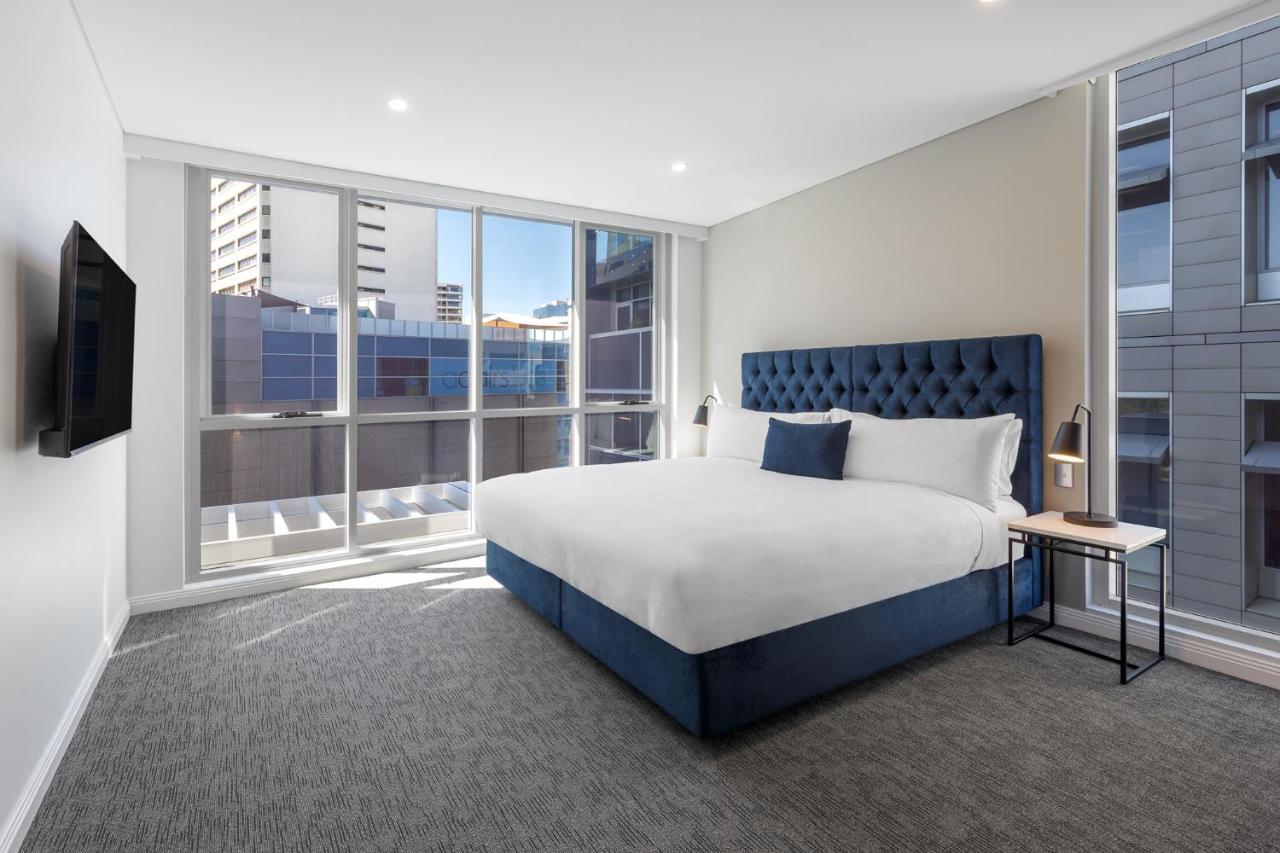 Meriton Suites Bondi Junction Sidney Exterior foto The image shows a modern hotel room with a large, comfortable bed featuring a blue upholstered headboard. The room has large windows that allow natural light to enter, providing a good view of the surrounding buildings. There is a small bedside table