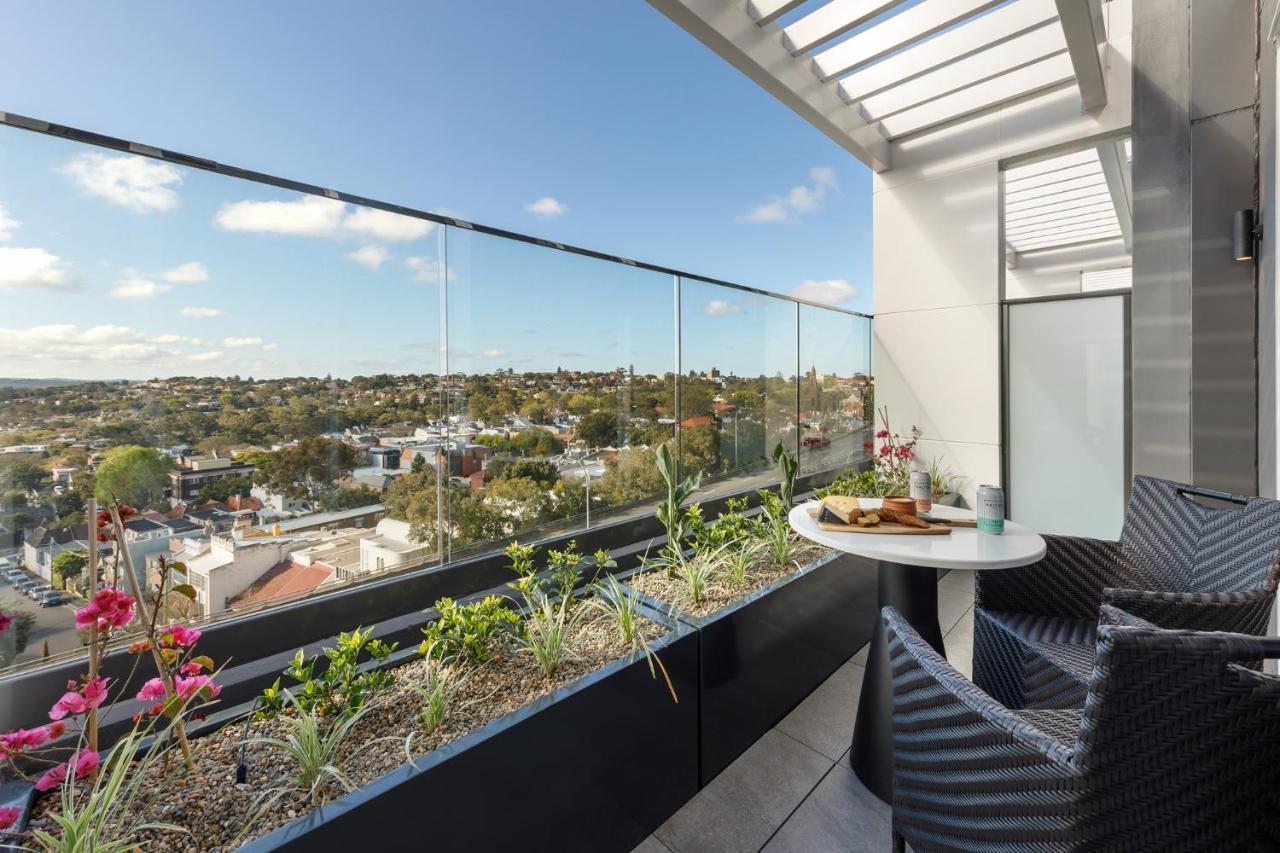 Meriton Suites Bondi Junction Sidney Exterior foto The photo shows a modern balcony with a glass railing, enhancing the view of the surrounding area, which includes various rooftops and greenery in the distance. There is a small round table set with a decorative floral arrangement and some items, lik