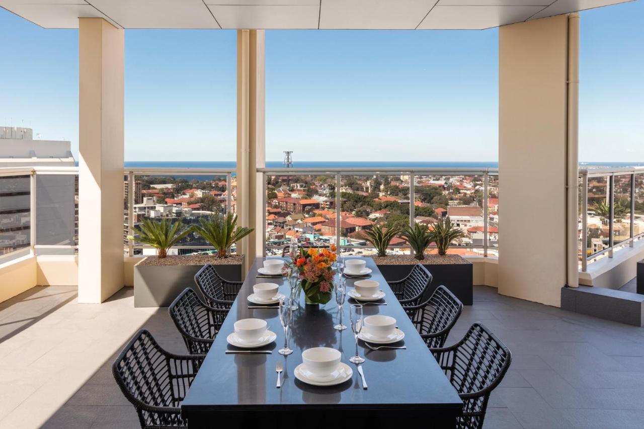 Meriton Suites Bondi Junction Sidney Exterior foto The photo shows a modern outdoor dining area situated on a balcony or terrace with a stunning view. There is a long rectangular table set with plates, glasses, and cutlery, ready for a meal. The table is surrounded by sleek, black wicker chairs. In t
