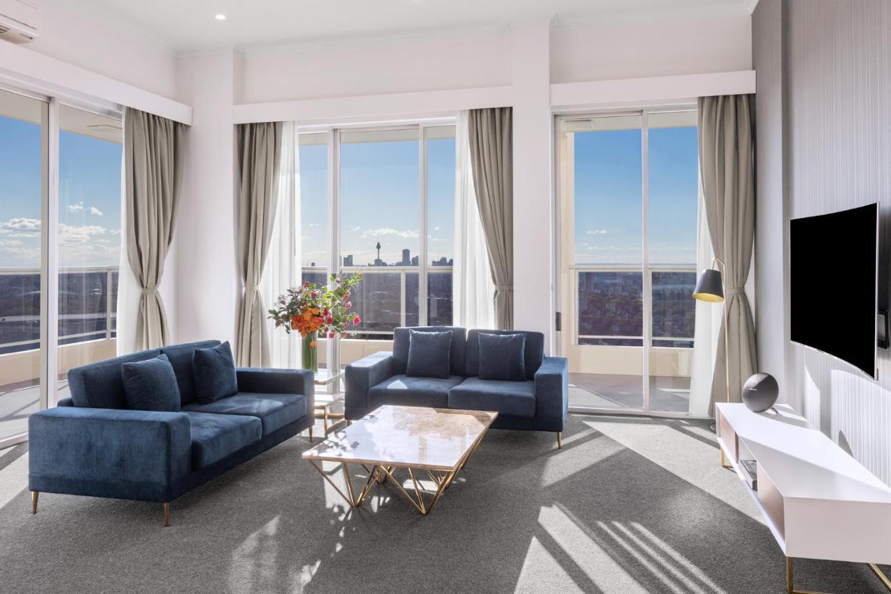 Meriton Suites Bondi Junction Sidney Exterior foto The photo shows a modern living room with large windows that allow natural light to fill the space. There are two blue sofas arranged around a coffee table with a glass top and gold legs. A floral arrangement adds a touch of color, placed on a nearby