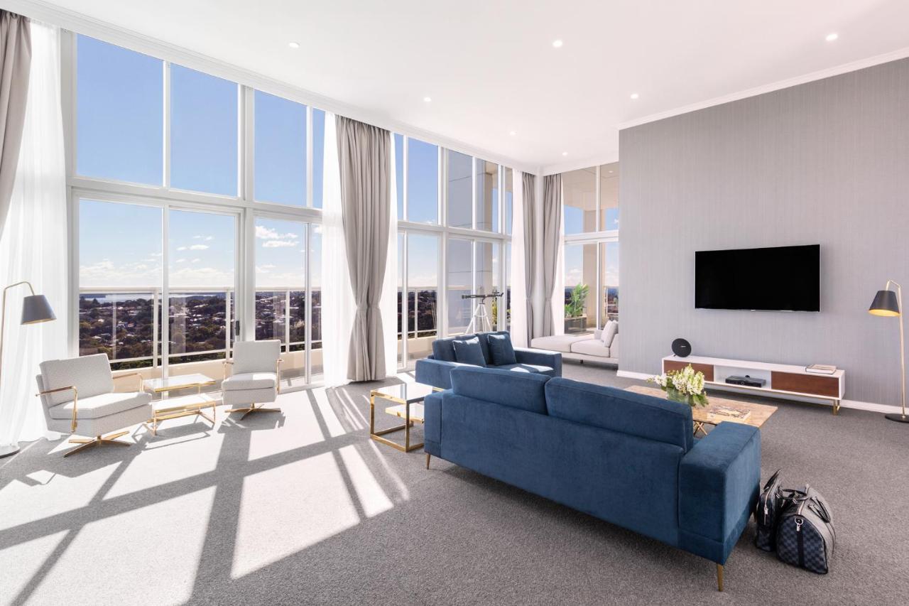 Meriton Suites Bondi Junction Sidney Exterior foto The photo shows a bright and modern living room with large floor-to-ceiling windows that provide a view of the outdoors. The space features a blue sofa in the center, complemented by light-colored armchairs nearby. There are light curtains hanging by
