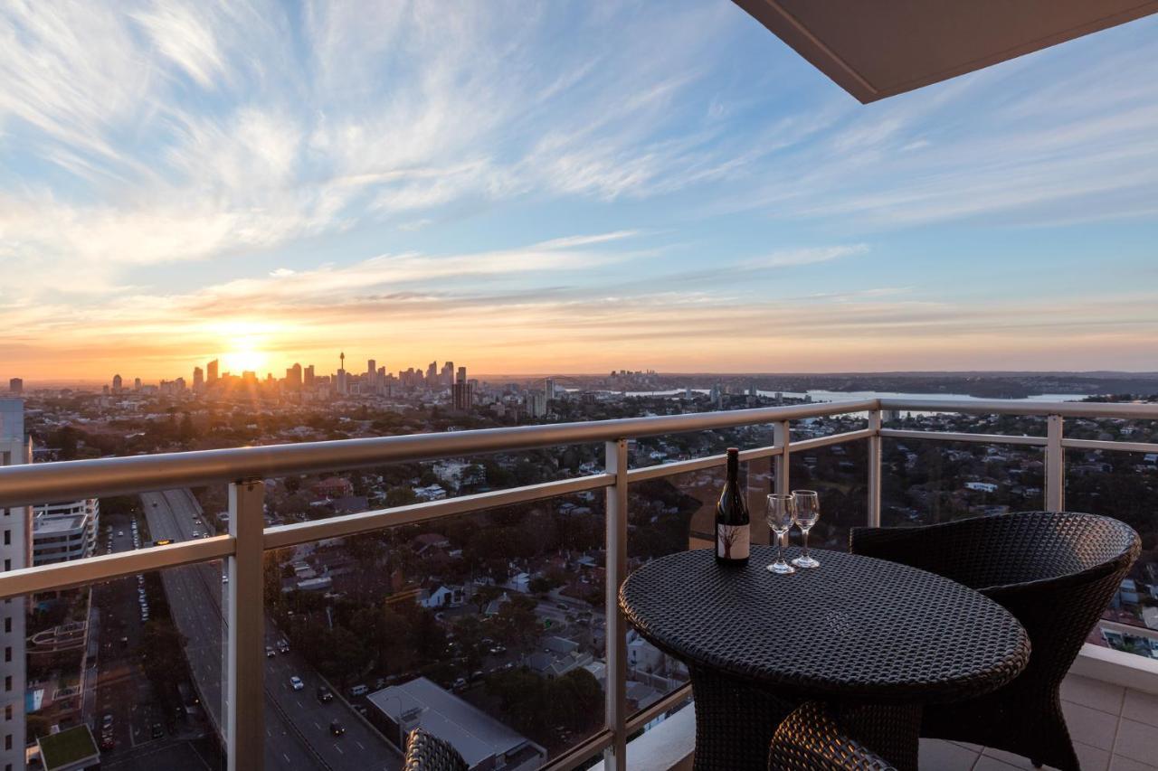 Meriton Suites Bondi Junction Sidney Exterior foto The image shows a scenic view from a balcony or terrace overlooking a cityscape during sunset. In the foreground, there is a small round table with two glasses and a bottle of wine, suggesting a romantic or relaxing setting. The sky is filled with vi