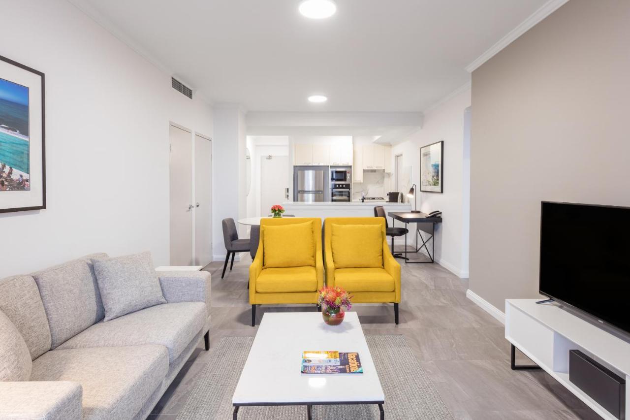 Meriton Suites Bondi Junction Sidney Exterior foto The photo shows a modern living space with a light and airy design. In the foreground, there are two bright yellow armchairs flanking a white coffee table, which has a small vase of flowers and a magazine on it. To the left, there is a light gray sof