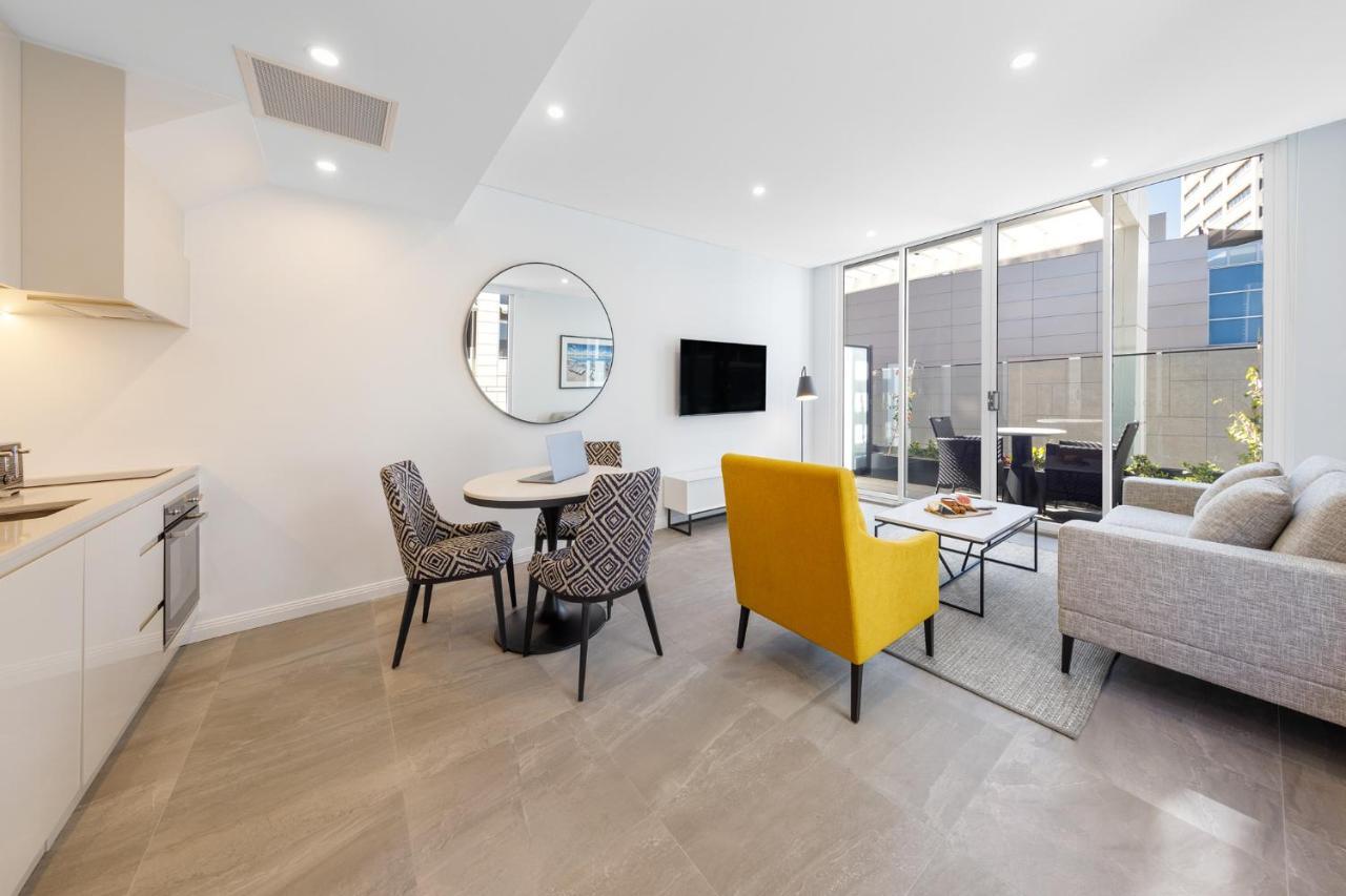Meriton Suites Bondi Junction Sidney Exterior foto The photo shows a modern and stylish living space. On one side, there is a kitchen area with sleek cabinetry, and next to it, a round dining table surrounded by three patterned chairs. In the center of the room, there is a gray sofa accompanied by a 