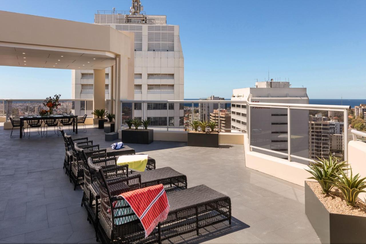 Meriton Suites Bondi Junction Sidney Exterior foto The photo shows a rooftop terrace with a modern design. There are several outdoor seating arrangements featuring black wicker lounge chairs. Some of the chairs have colorful blankets draped over them. The terrace is surrounded by a railing and has po
