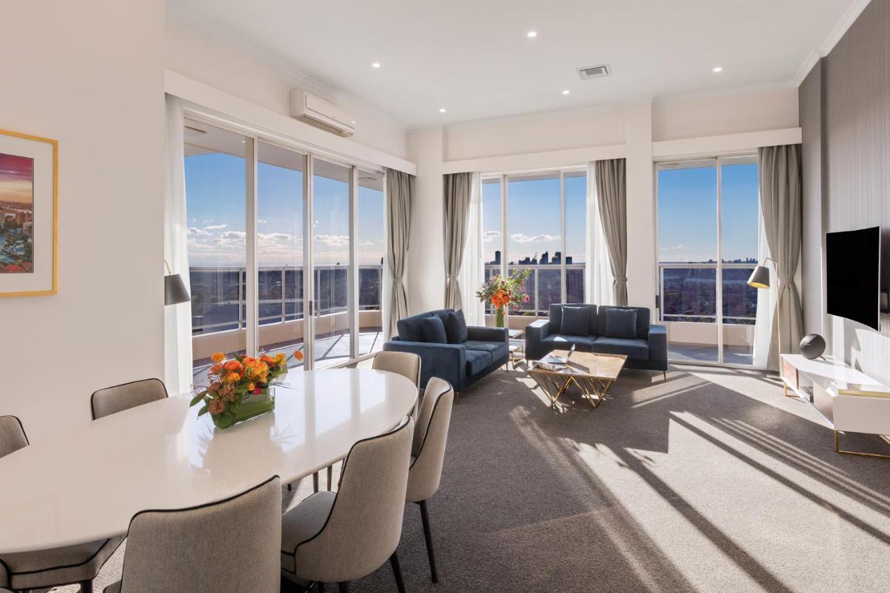 Meriton Suites Bondi Junction Sidney Exterior foto The photo shows a modern and stylish living space with large windows allowing natural light to fill the room. In the foreground, there is a round dining table with several chairs around it, adorned with a floral centerpiece. The seating area features