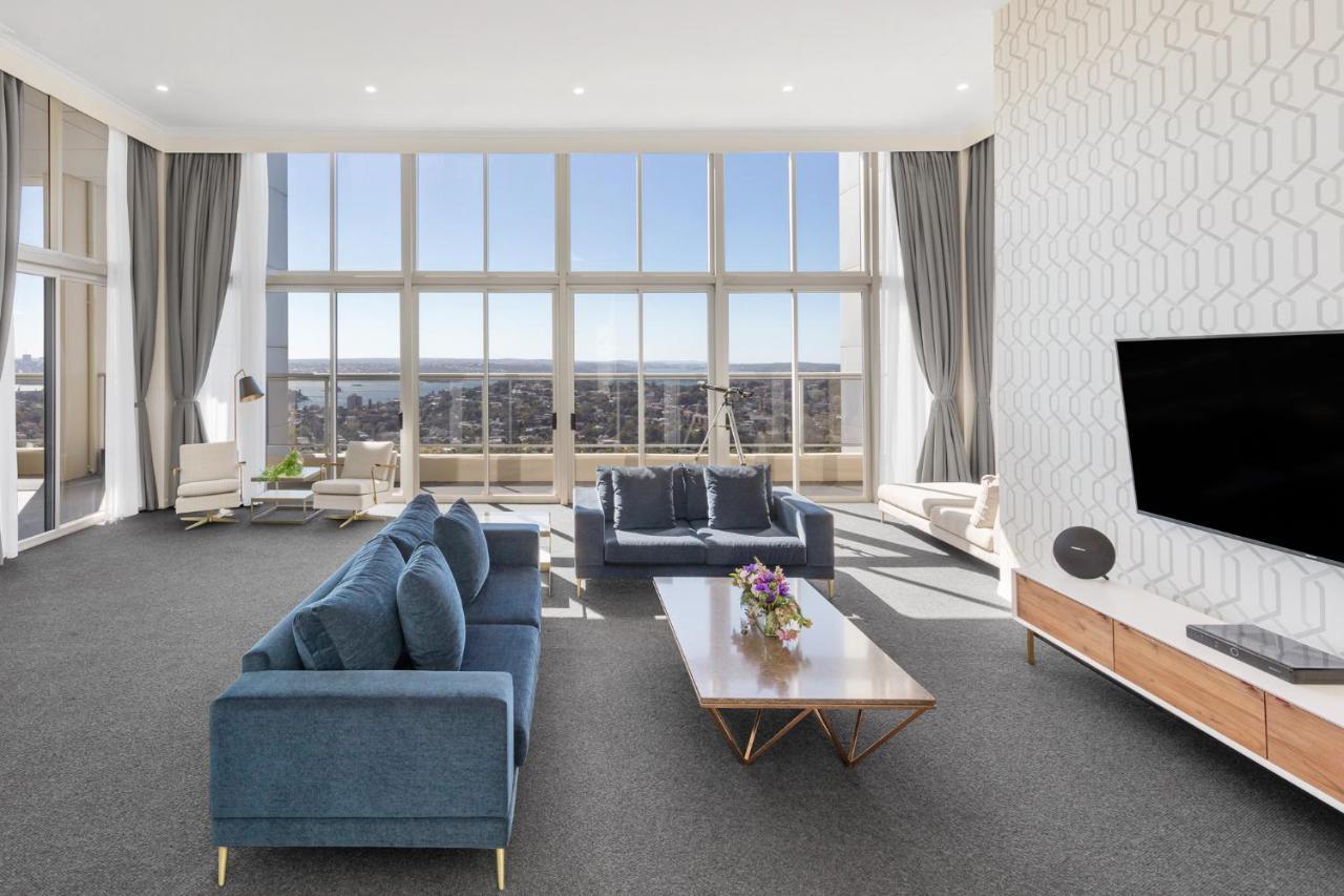 Meriton Suites Bondi Junction Sidney Exterior foto The photo shows a modern and spacious living area. It features large windows that provide a panoramic view of the outside landscape and allow natural light to fill the room. The interior design includes a blue sectional sofa placed in the center, com