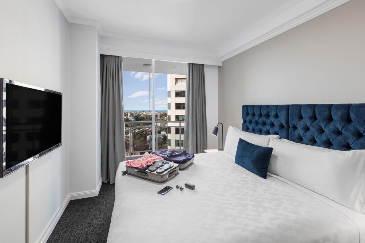 Meriton Suites Bondi Junction Sidney Exterior foto The photo shows a modern hotel room featuring a neatly made bed with a blue tufted headboard and a white duvet. There's a small decorative pillow on the bed, and a piece of luggage is placed at the foot with clothes partially unpacked. A television i