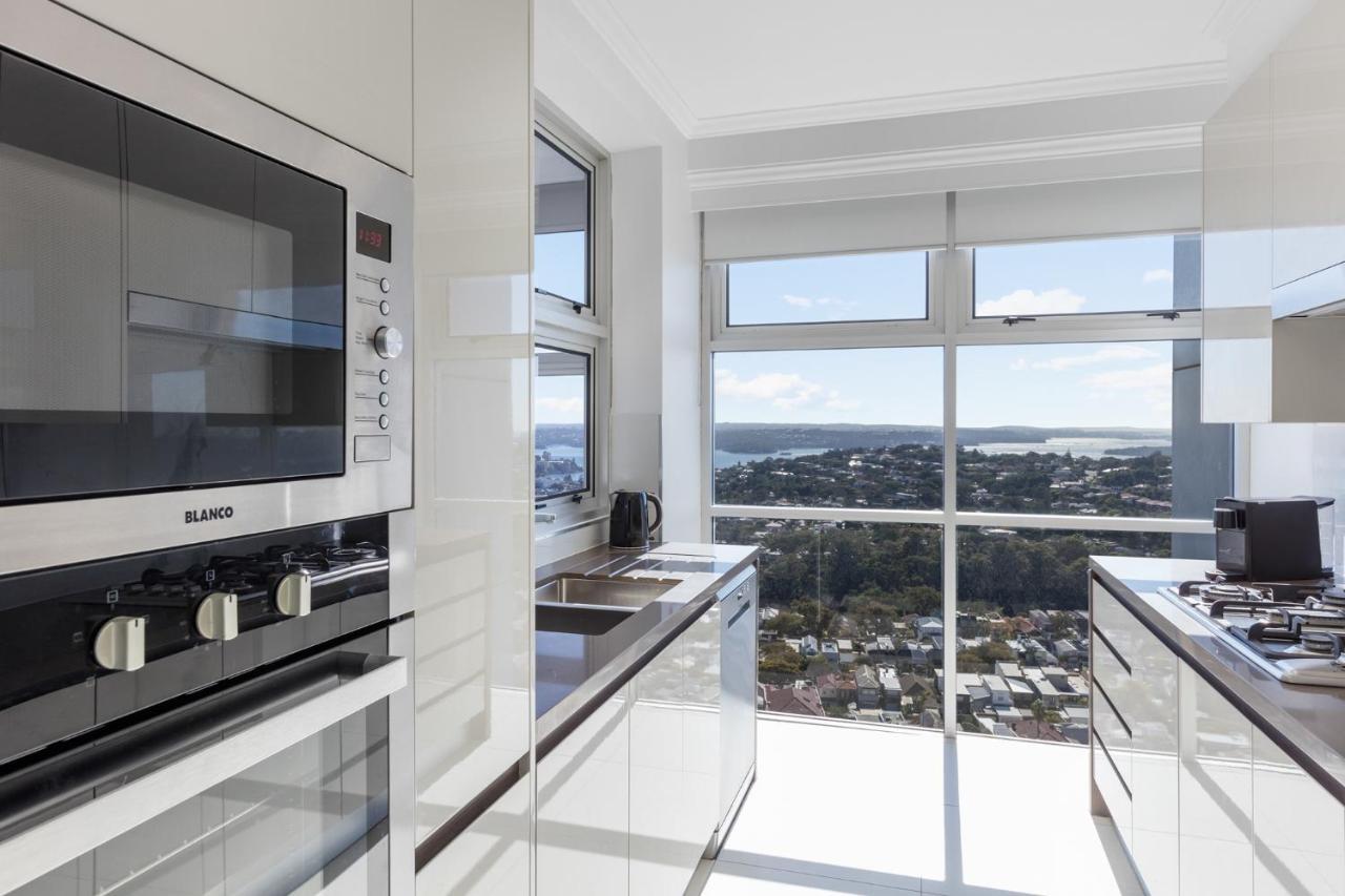 Meriton Suites Bondi Junction Sidney Exterior foto The photo shows a modern kitchen with sleek, shiny white cabinets and a large window that offers a panoramic view of the outside landscape. The kitchen features built-in appliances, including an oven and a stove, with a stylish countertop. The spacio