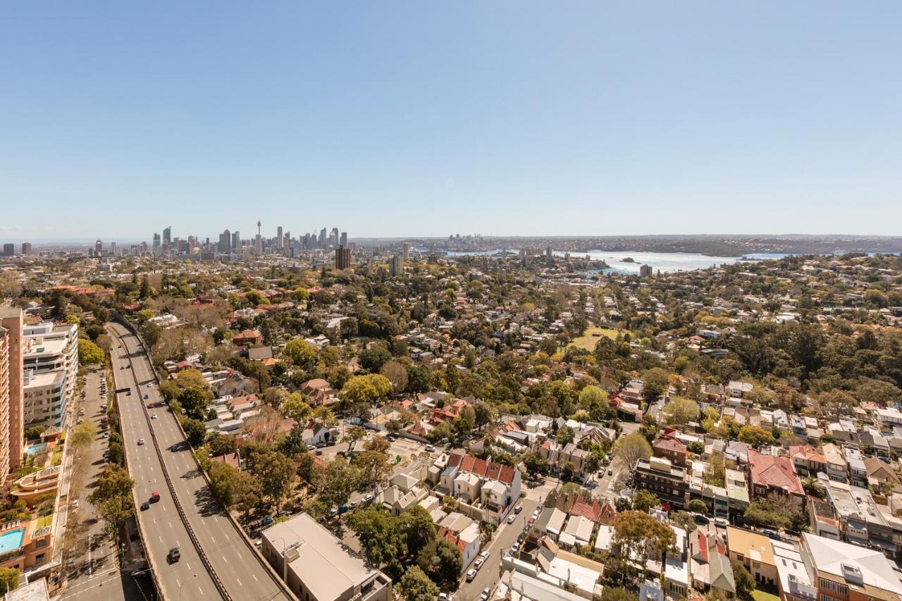 Meriton Suites Bondi Junction Sidney Exterior foto The photo captures a panoramic view of a vibrant urban landscape. In the foreground, there are residential areas with various houses and greenery, showcasing a suburban environment. In the background, the skyline of a city can be seen, featuring tall