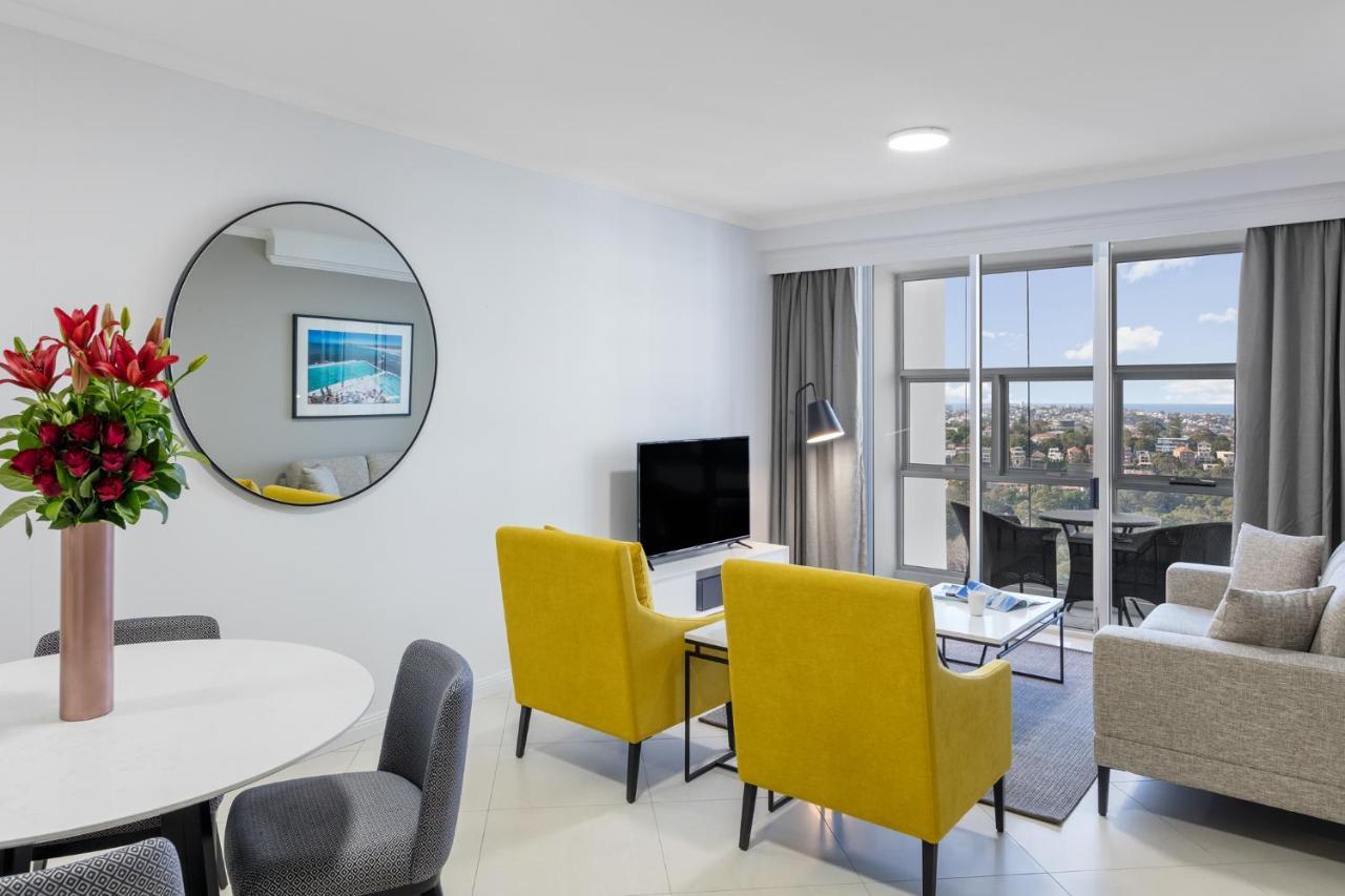 Meriton Suites Bondi Junction Sidney Exterior foto The photo shows a modern living space with a stylish design. There are two bright yellow chairs facing a small glass table, and a comfortable beige sofa to the right. A TV is positioned on the wall in front of the chairs. To the left, there's a dinin