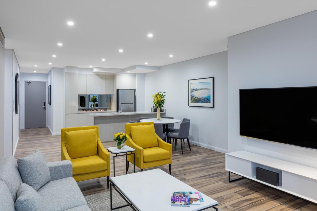 Meriton Suites Bondi Junction Sidney Exterior foto The photo shows a modern living space that includes a cozy sitting area and a dining area. The sitting area features two bright yellow chairs and a light gray sofa, arranged around a coffee table. There are decorative items on the table, including fl