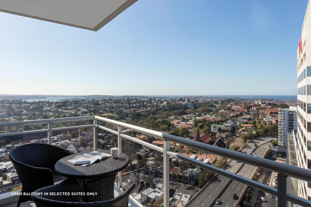 Meriton Suites Bondi Junction Sidney Exterior foto The photo shows a balcony with a view over a cityscape that includes buildings and greenery, extending towards the horizon where the sea is visible. The balcony features a small round table and two black chairs, suggesting a space for relaxation or d