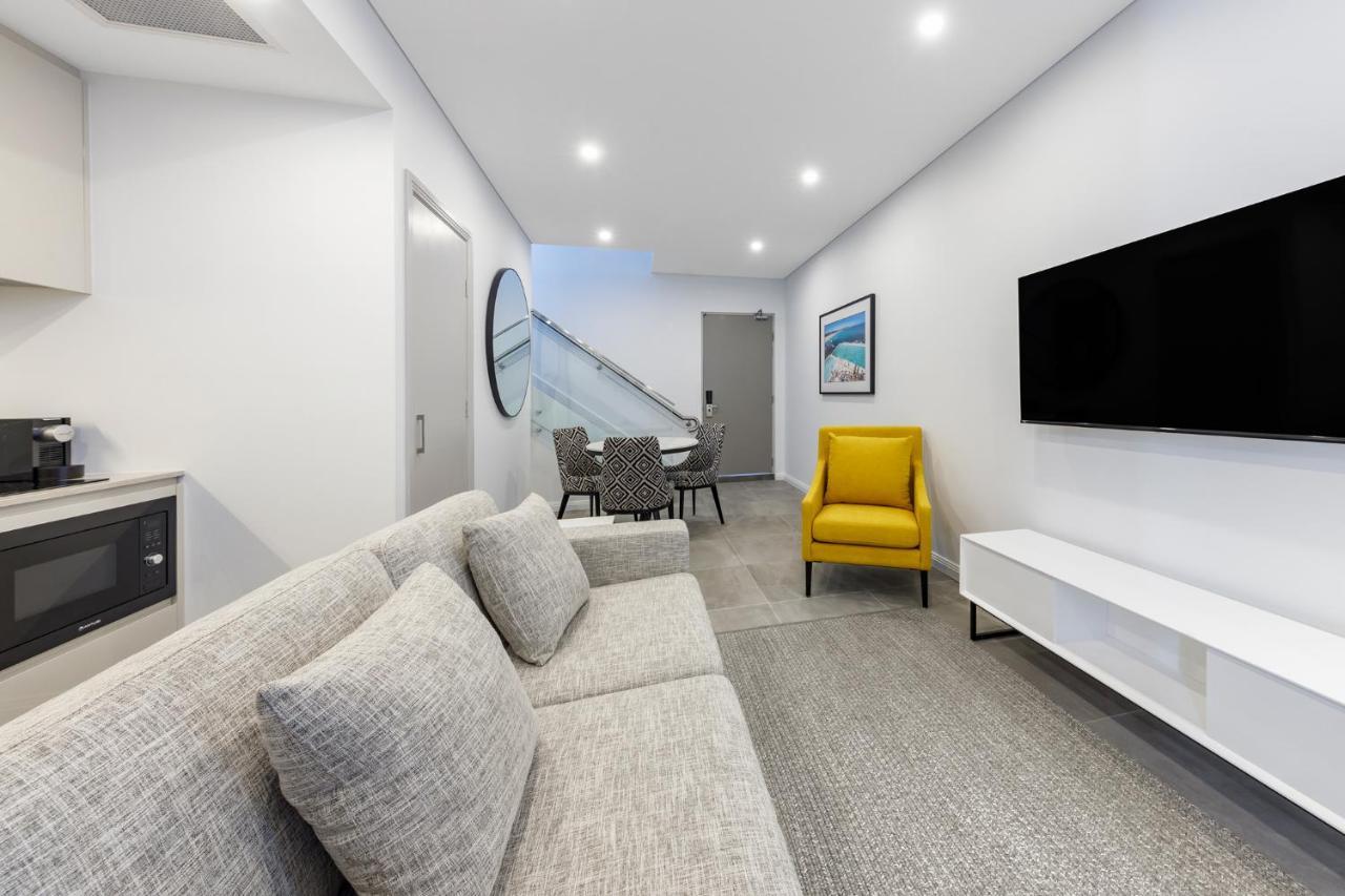 Meriton Suites Bondi Junction Sidney Exterior foto The photo shows a modern and stylish living space. It features a light-colored sofa with cushions, positioned to face a wall-mounted flat-screen TV. There's a yellow accent chair nearby that adds a pop of color to the room. The décor is minimalistic,