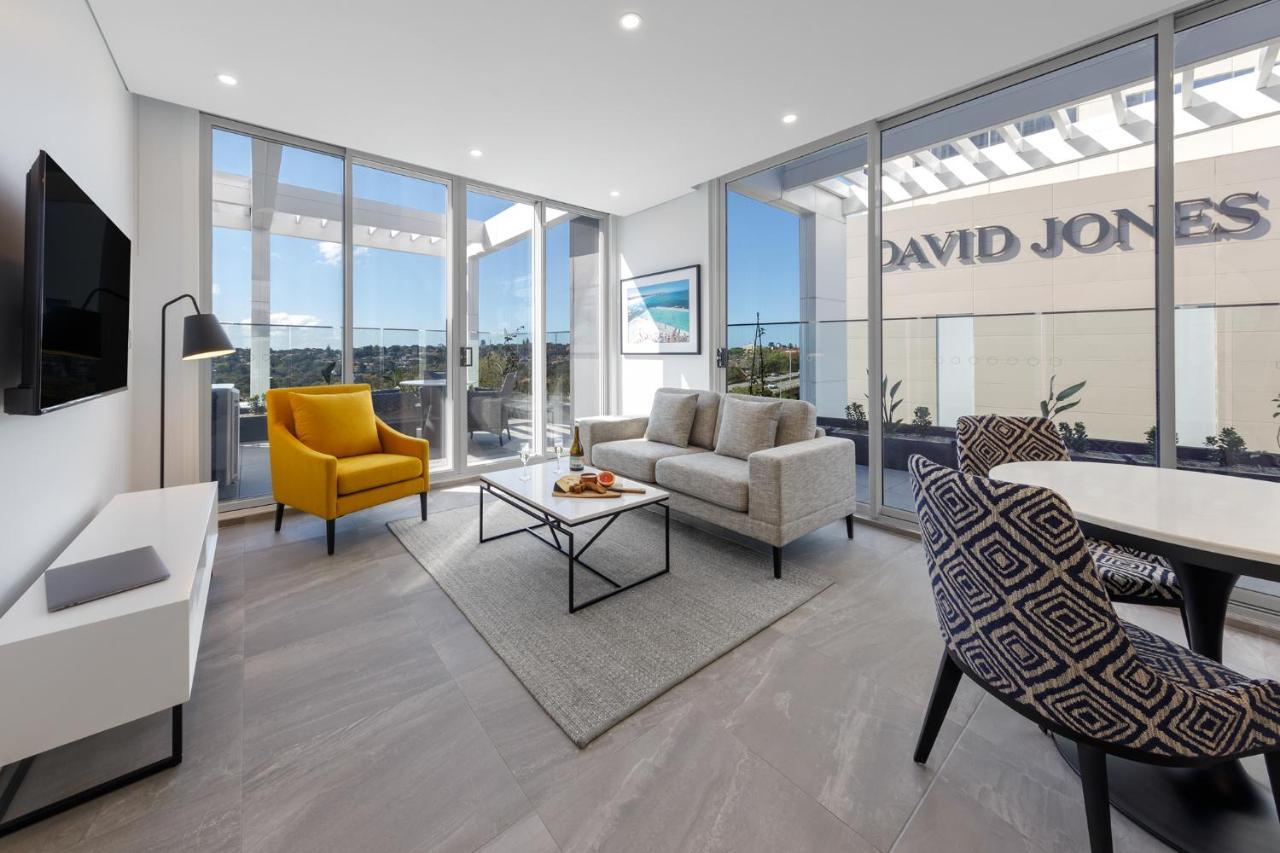 Meriton Suites Bondi Junction Sidney Exterior foto The photo shows a modern living space that features large windows allowing ample natural light. The room is furnished with a gray sofa and a bright yellow armchair, along with a coffee table in between. There’s a decorative bowl with fruits on the ta