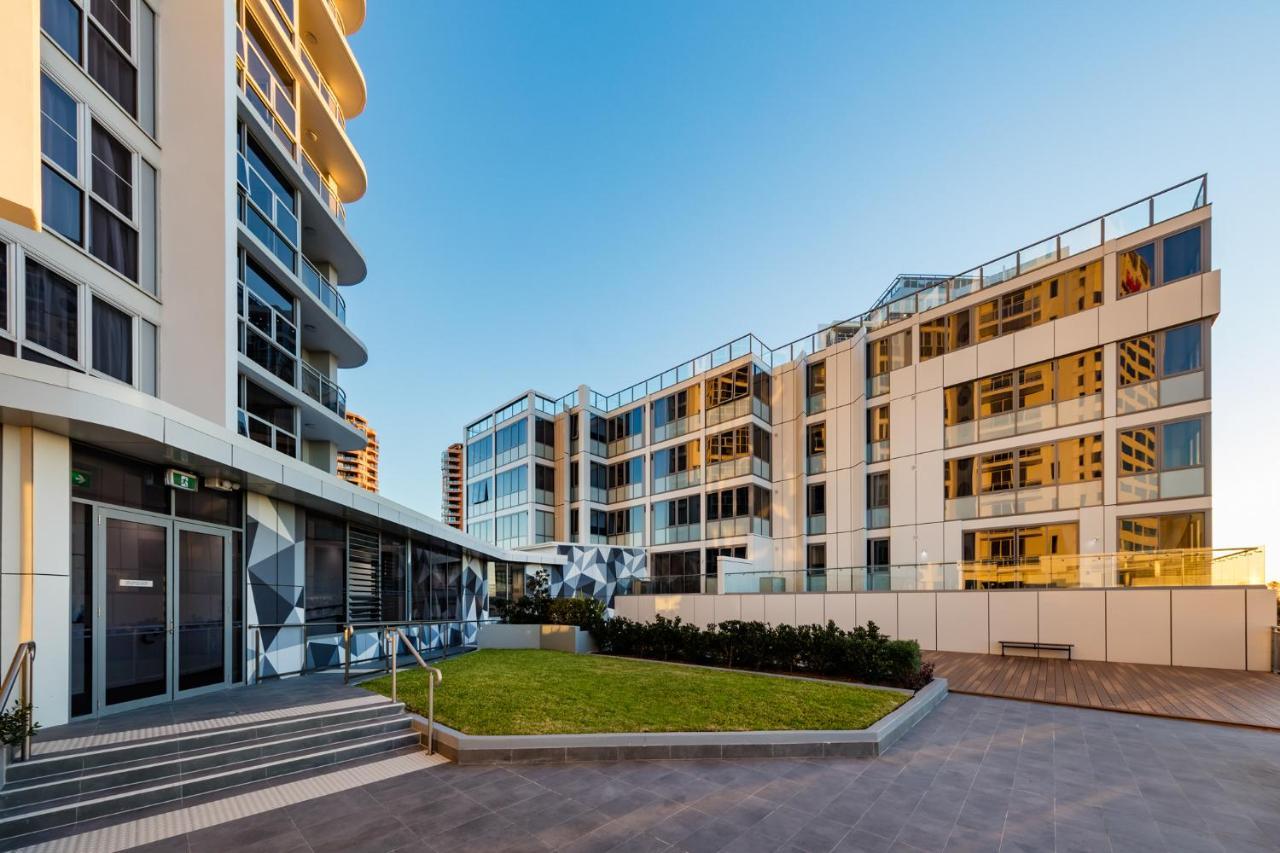 Meriton Suites Bondi Junction Sidney Exterior foto The photo showcases a modern residential area featuring contemporary buildings. In the foreground, there is a landscaped green space surrounded by well-designed architecture. The buildings have large glass windows, reflecting the light, and feature a