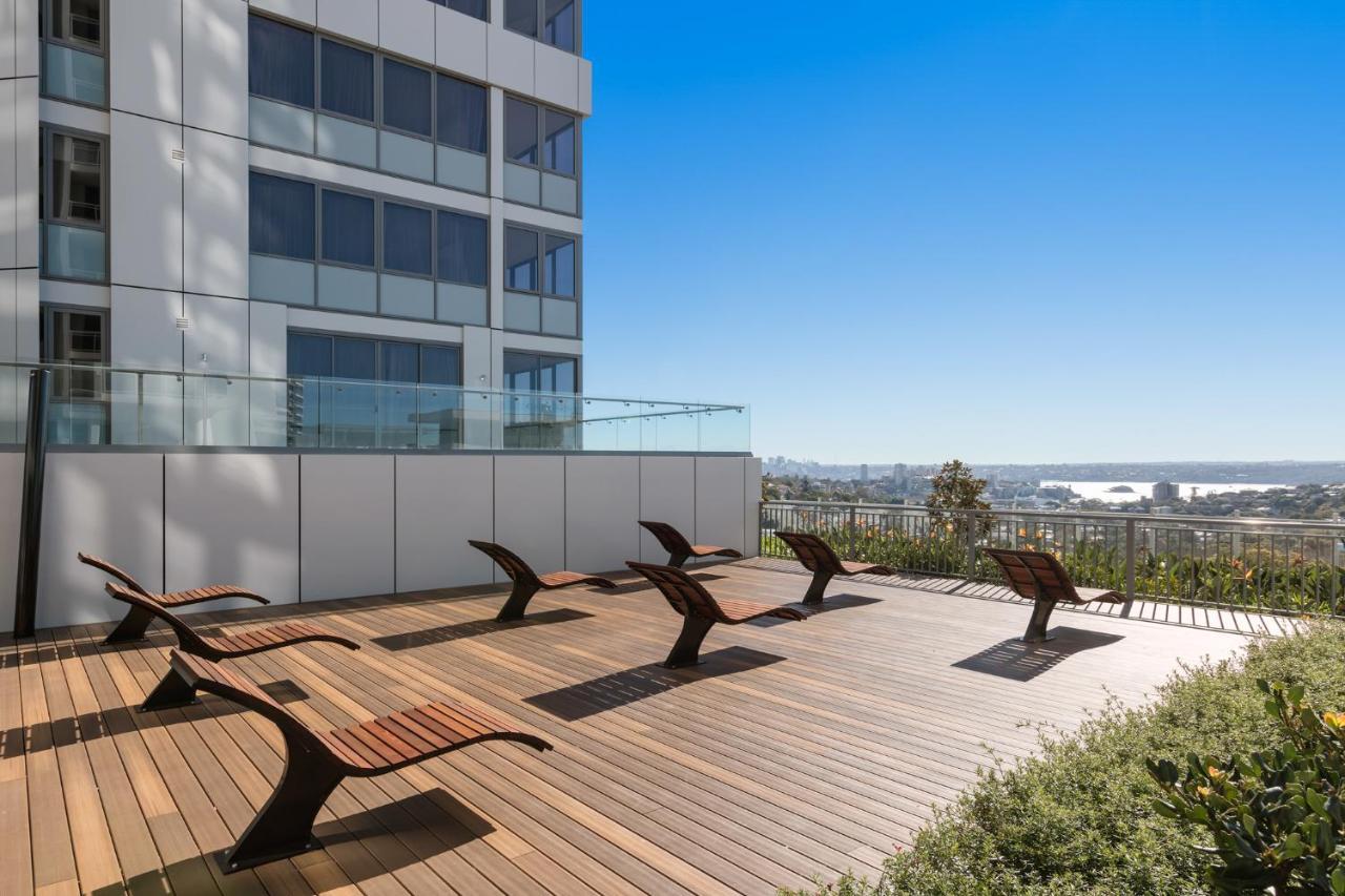 Meriton Suites Bondi Junction Sidney Exterior foto The photo shows a rooftop or outdoor area with a wooden deck. There are several modern lounge chairs arranged neatly, facing a clear sky and a panoramic view of the surrounding landscape. In the background, there is a tall building with large windows