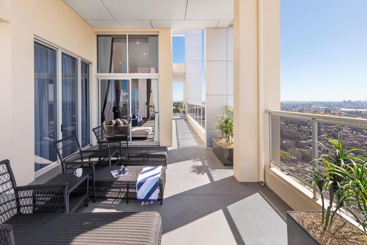 Meriton Suites Bondi Junction Sidney Exterior foto The photo shows a spacious balcony or terrace area of a high-rise building. It features modern outdoor furniture, including a seating arrangement with dark wicker chairs and a table. There are cushions, and a blue and white striped blanket is draped 