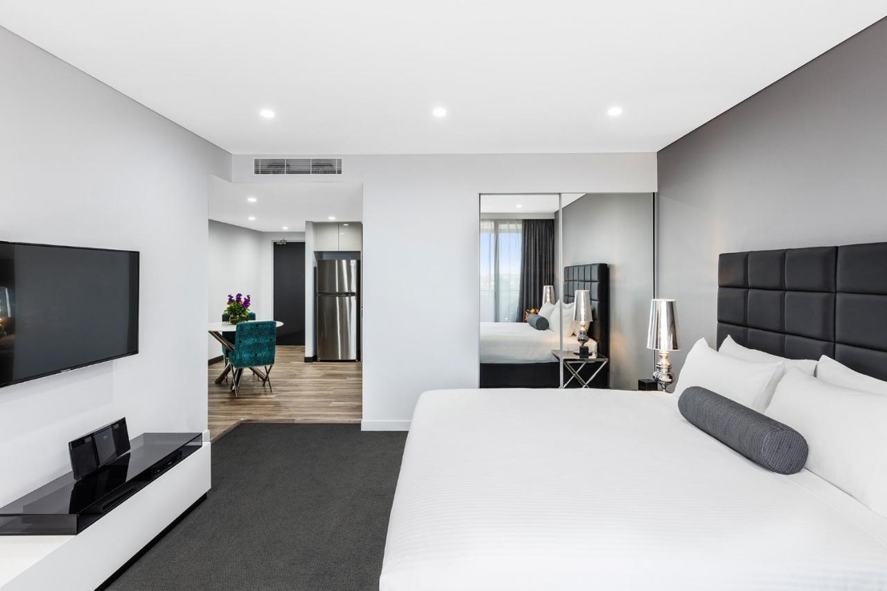 Meriton Suites Bondi Junction Sidney Exterior foto The photo shows a modern hotel room. It features a large bed with a white covering and gray accent pillows. To the left, there's a flat-screen TV mounted on the wall above a sleek white cabinet. The flooring is a light wood tone, and a large mirror i
