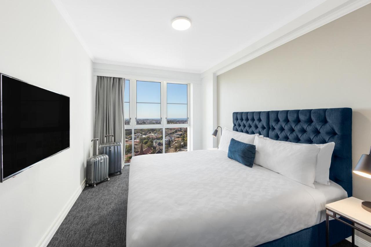 Meriton Suites Bondi Junction Sidney Exterior foto The photo shows a modern hotel room with a large bed featuring a tufted blue headboard. There is a white duvet and a decorative pillow on the bed. A flat-screen television is mounted on the wall opposite the bed. Large windows are covered with gray c