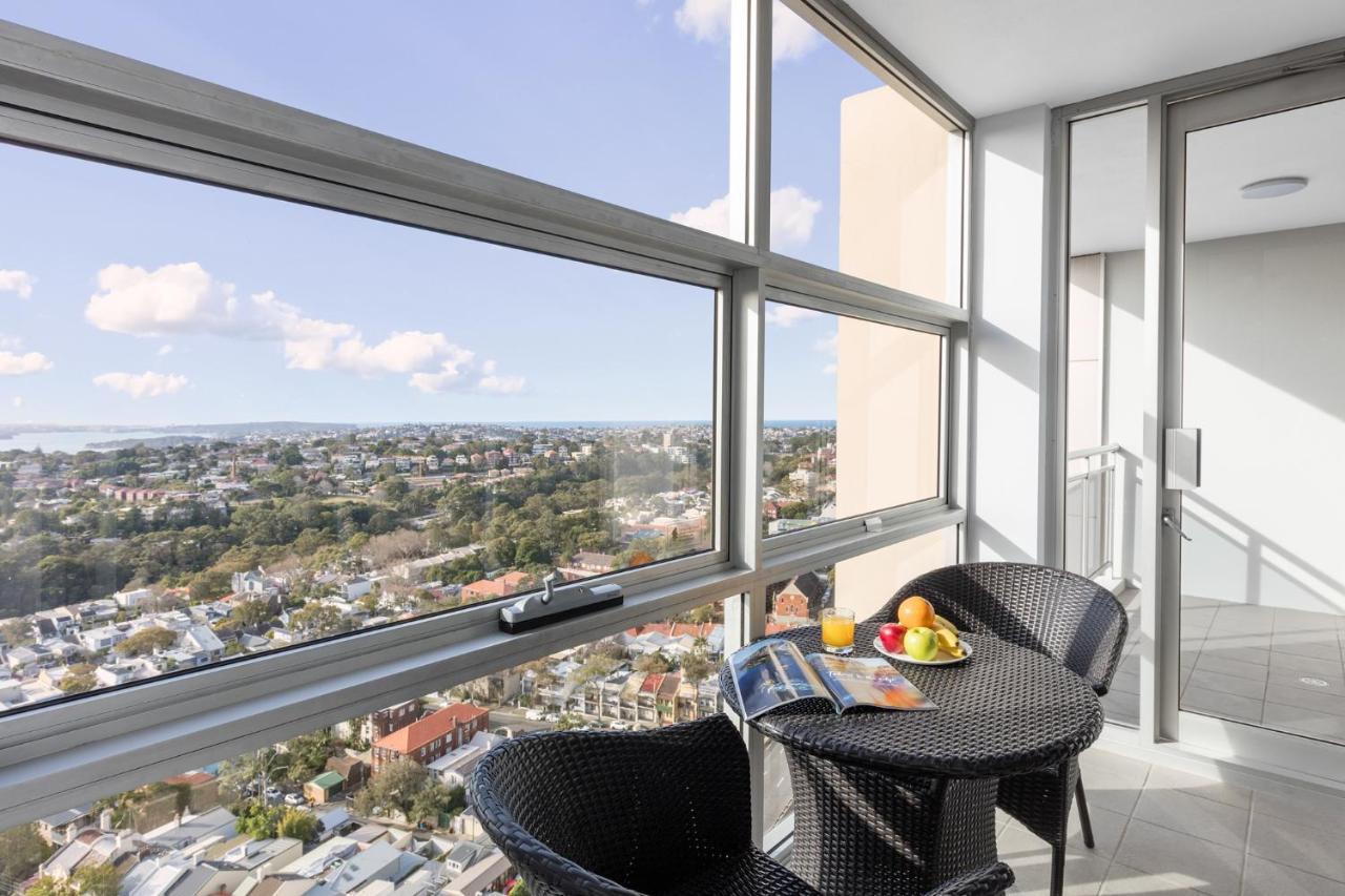 Meriton Suites Bondi Junction Sidney Exterior foto The photo shows a modern balcony with large glass windows that offer a panoramic view of a sprawling urban landscape. There is a small round table made of dark wicker, accompanied by two matching chairs. On the table, there is a bowl of fruit and a m