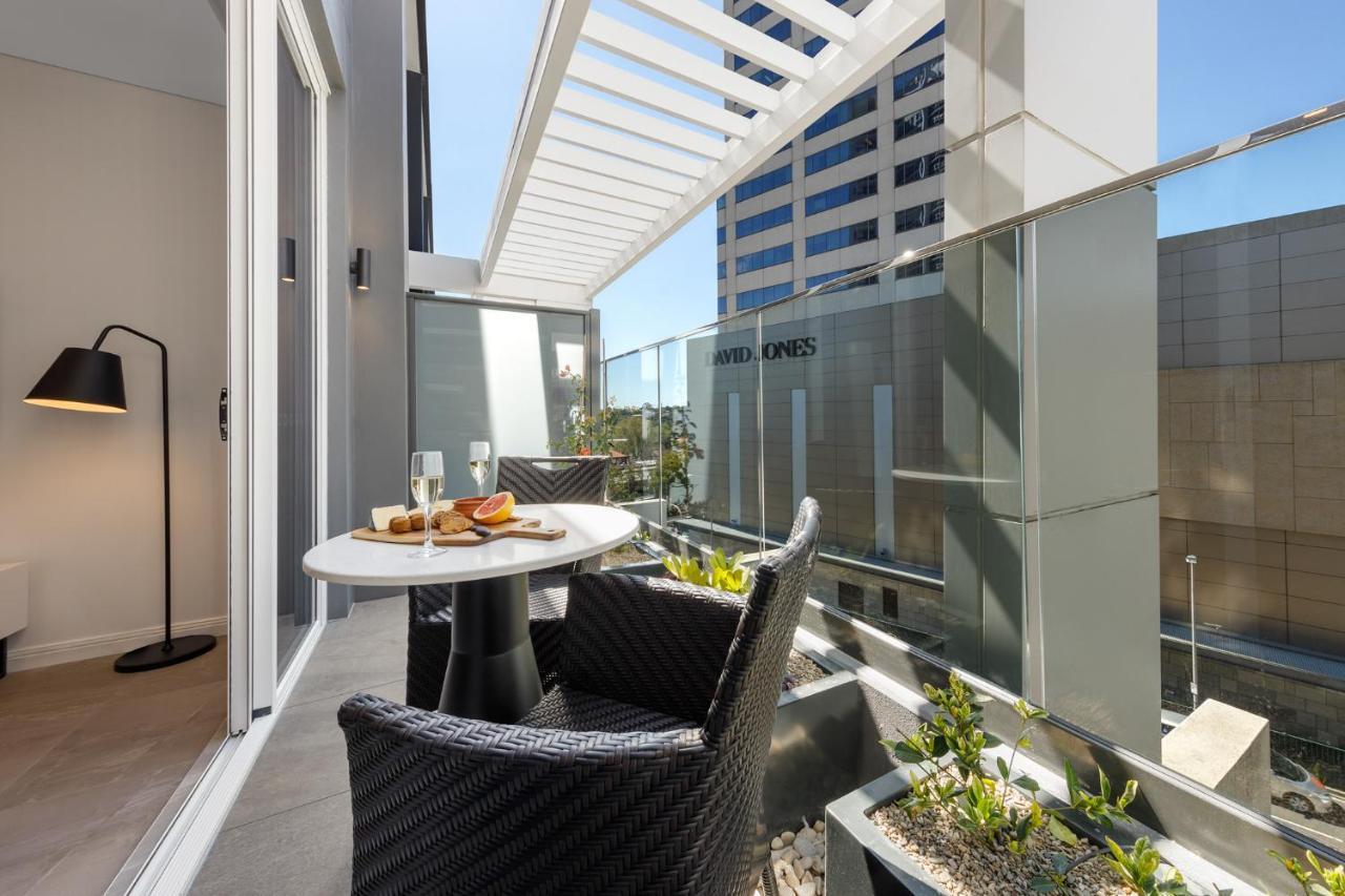 Meriton Suites Bondi Junction Sidney Exterior foto The photo shows a modern balcony area with glass railings. It features a small round table set for two, with a plate of food and drinks. There are two stylish wicker chairs positioned around the table. The space is bright and airy, with sunlight stre