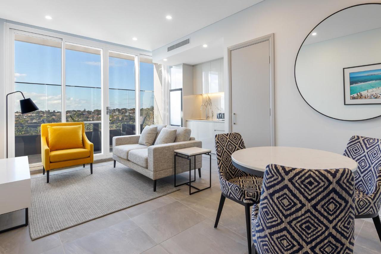 Meriton Suites Bondi Junction Sidney Exterior foto The photo shows a modern and stylish living area. It features a comfortable sofa in a neutral color, accompanied by a bright yellow armchair that adds a pop of color. There's a round dining table with patterned chairs nearby. Large windows allow plen