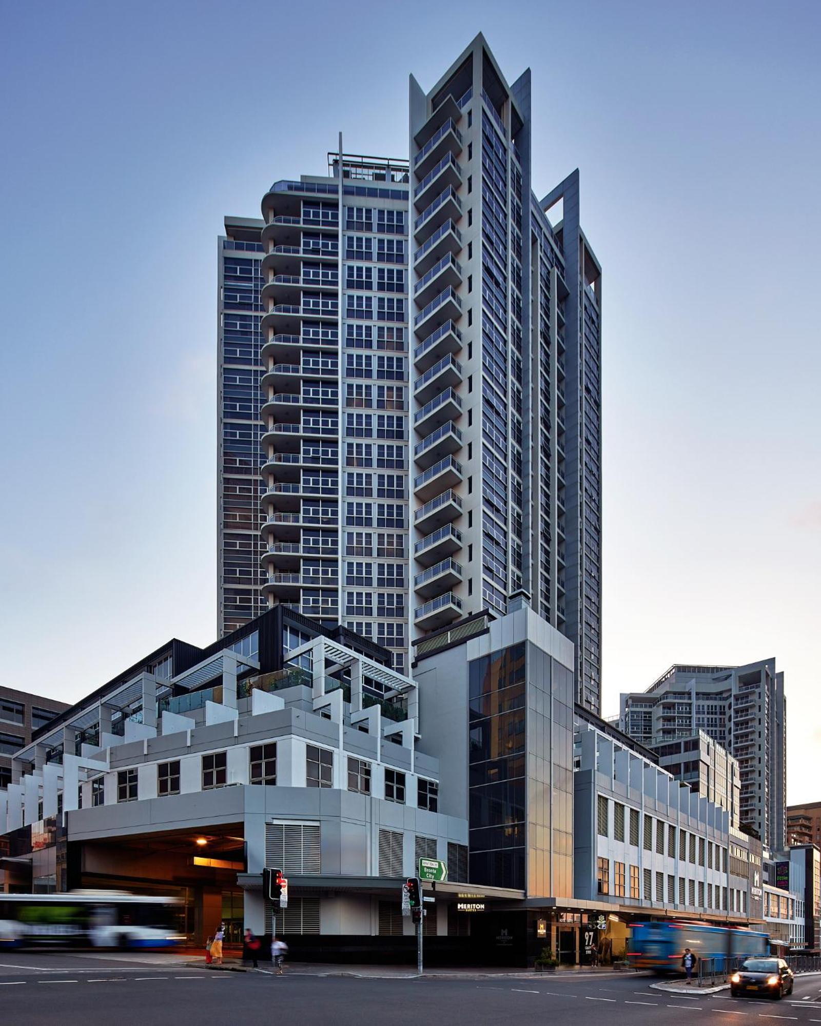 Meriton Suites Bondi Junction Sidney Exterior foto The image shows a modern high-rise building with a sleek design. The structure features a mix of glass and concrete elements, with multiple levels showcasing balconies. The building appears to be part of an urban environment, likely containing reside