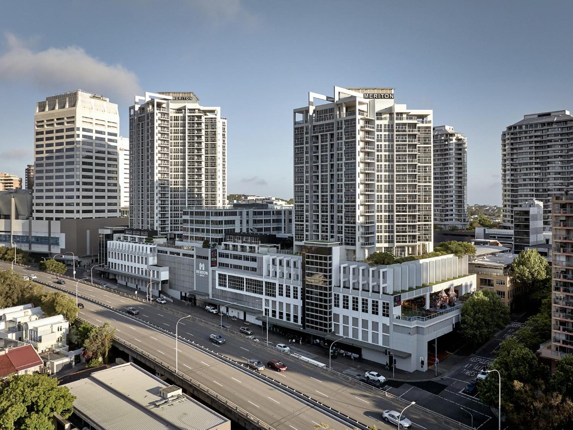 Meriton Suites Bondi Junction Sidney Exterior foto The photo shows a cityscape featuring several tall modern buildings, likely apartments or office complexes. In the foreground, there is a road lined with cars, and the architecture of the buildings varies, with some having sleek glass facades and oth