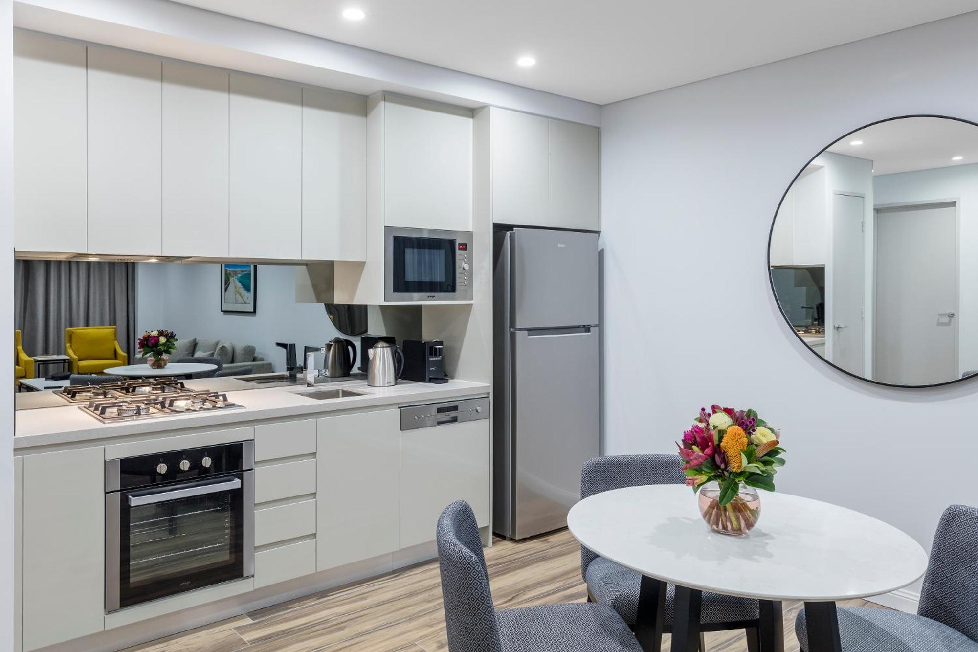Meriton Suites Bondi Junction Sidney Exterior foto The photo shows a modern kitchen and dining area. The kitchen features light-colored cabinetry with a built-in oven and stovetop, a microwave, and a silver refrigerator. There is a kettle and a coffee maker on the countertop. In the dining area, ther