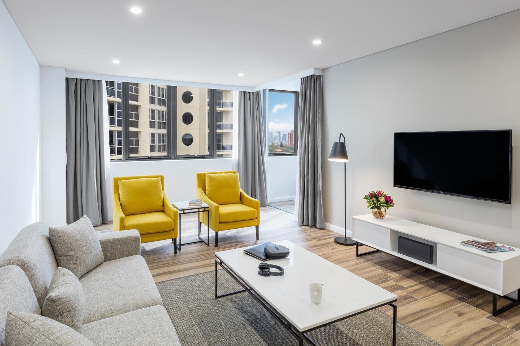 Meriton Suites Bondi Junction Sidney Exterior foto The photo shows a modern, well-lit living room. It features a light gray sofa with a textured fabric. There are two bright yellow armchairs positioned near a window, which allows natural light to enter the space. The room has light gray curtains and 